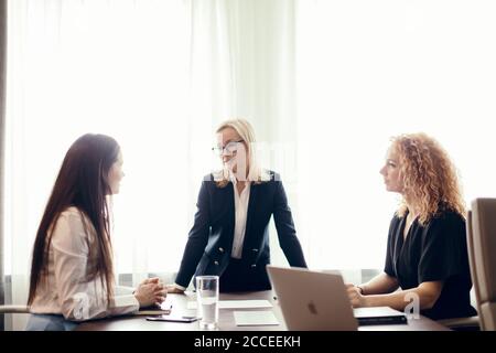 Group of business women meeting at the office. Blonde woman dressed in formal suit sharing her ideas with two colleagues, sitting at the desk. Stock Photo