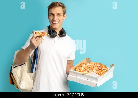 portrait of young caucasian pizza lover isolated over blue background, male in white t-shirt with headphones hold pizza in hands, enjoy eating, delive Stock Photo