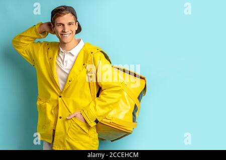 portrait of good-looking deliverman in special yellow uniform, carrying orders in backpack for customers and looking at camera isolated over blue back Stock Photo