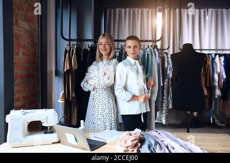 Two happy women look at camera in workplace. Beautiful caucasian woman in dress and pretty female designer in shirt. Laptop, sewing machne on table. B Stock Photo