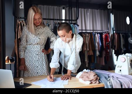 Modern designer and cheerful client discuss and project new dress in workplace, on table with papers and laptop. Stock Photo