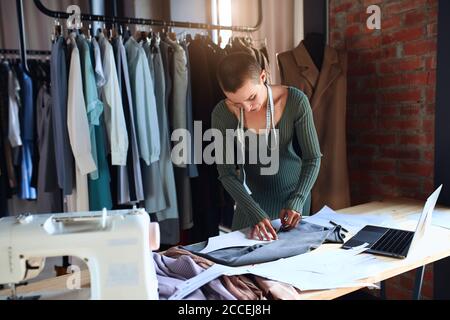 Female dressmaker sew new clothing in studio using laptop. Background rack of clothes Stock Photo