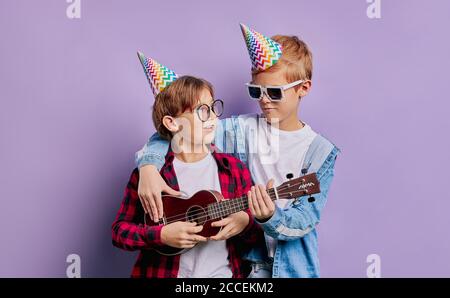 two happy kids friends hugging celebrate birtday of one of them, wearing eyeglasses and birthday hats, boy in red shirt holding ukulele in hands, smal Stock Photo