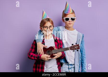 portrait of beautiful kid boys at happy birthday wearing holiday hat on head and holding ukulele, performing music. friendship, children, birthday con