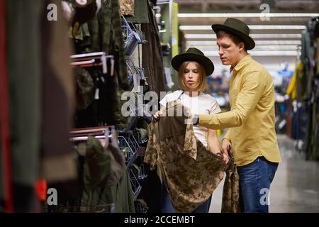 beautiful married couple going to go in mountains, shopping together in sportive wear store, handsome guy and beautiful woman try on green wear and ha Stock Photo