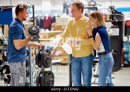 young affable consultant of sportswear shop talks with customers, show the best variant of dumbbells, rise it with strong muscular hands, customers ey Stock Photo