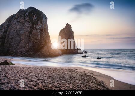 Sunset at Ursa Beach Sea stack, Portugal. Atlantic Ocean Foamy waves rolling to sandy beach. Holiday vacation landscape scene. Stock Photo