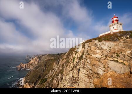 Cabo da Roca Lighthouse at cliff edge in evening sunlight against beautiful cloudscape. Most westerly point of the Europe mainland, Sintra, Portugal. Stock Photo