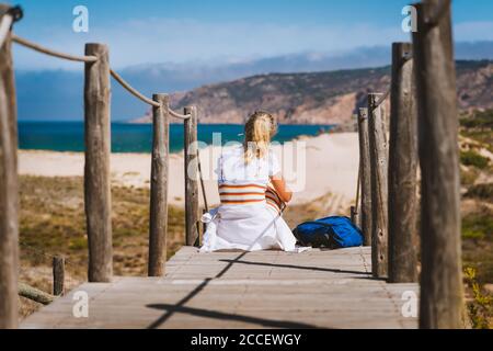 Adult female tourist sitting on path leading to Praia do Guincho Beach. Cascais, Portugal. This is popular blue flag Atlantic beach for surfing, winds Stock Photo
