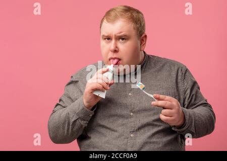 Morning routine concept. Suspicious sceptical man holds tooth brush in hand, taking toothpaste on taste, wears casual grey shirt, isolated on pink. Go Stock Photo