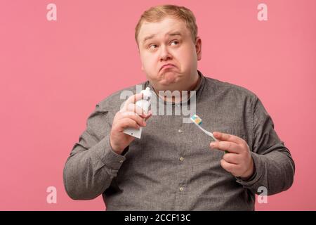 Questioning doubtful man with curved lips looking aside while holding toothbrush for cleaning teeth in hands, posing isolated over pink background. Stock Photo