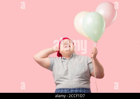 Middle aged happy funny man dressed in childish stylelaughin joyfully, holding colorful balloons prepares to celebrate birthday party of his son, pink Stock Photo