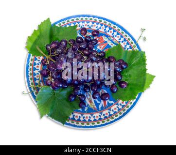 Top view looking down on fresh organic black grapes and grape leaves on a decorative colorful plate, isolated on a white background. Clipping path. Stock Photo