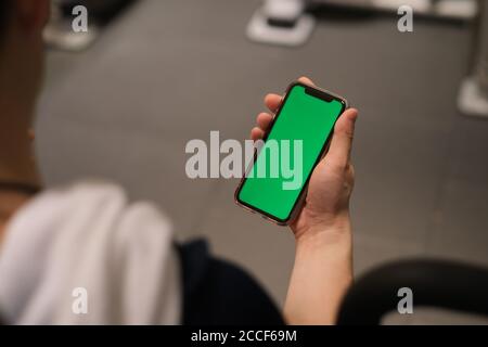 over shoulder of man holding green screen smart phone in the gym. blur background Stock Photo