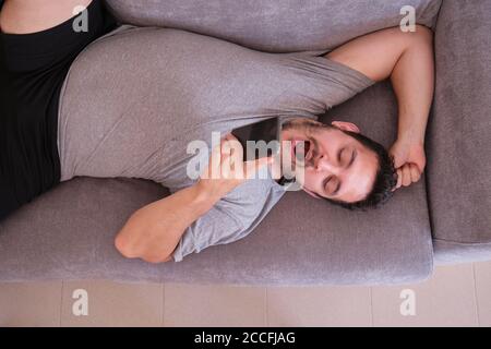 Top view of a young man yawning and looking something on his smartphone lying on couch at home. Technology concept. Stock Photo