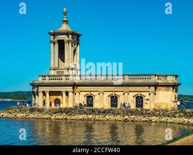 Historic Normanton Church standing at the end of a causeway on Rutland Water, with visitors outside in the sunshine against a blue sky. England, UK. Stock Photo