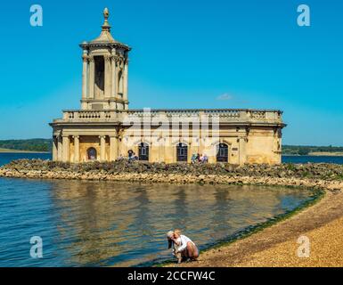 Historic Normanton Church standing at the end of a causeway on Rutland Water, with visitors outside in the sunshine against a blue sky. England, UK. Stock Photo