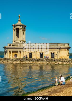 Historic Normanton Church standing at the end of a causeway on Rutland Water, with visitors outside in the sunshine against a blue sky. England, UK. Stock Photo