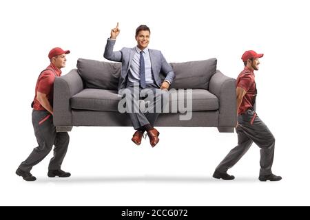 Smiling man in elegant clothes sitting on a sofa and pointing up while two movers are carrying the sofa isolated on white background Stock Photo