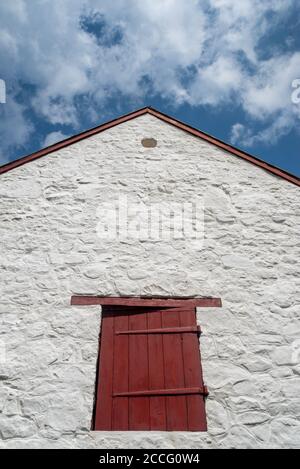 Whitewashed stone historic barn facade and red shutter under a blue sky. Long antique hinges and rough texture in natural light with copy space. Stock Photo