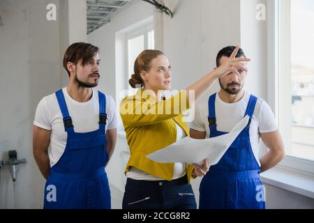 A client with a plan in her hands is talking to two construction workers in blue overalls Stock Photo