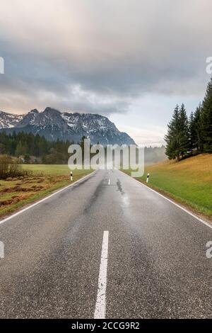 Early morning on the country road at Schmalensee near Mittenwald, the road is wet with rain, light fog rises and the Karwendel towers in the backgroun Stock Photo