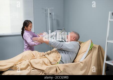 A little girl and her grandpa are happy to see each other, they are hugging, she came to see him at the hospital, he is staying in bed. Stock Photo