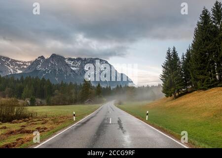 Early morning on the country road at Schmalensee near Mittenwald, the road is wet with rain, light fog rises and the Karwendel towers in the backgroun Stock Photo