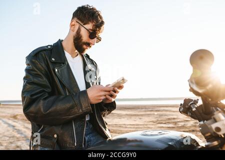 Handsome young bearded man wearing leather jacket sitting on a motorbike at the beach, using mobile phone Stock Photo