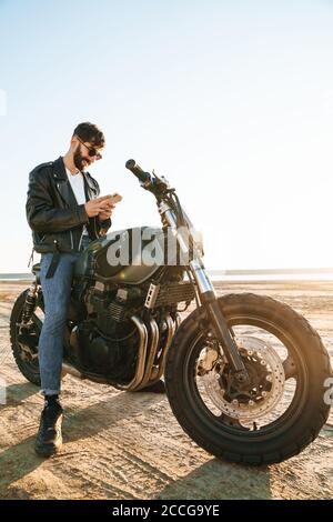 Handsome young bearded man wearing leather jacket sitting on a motorbike at the beach, using mobile phone Stock Photo