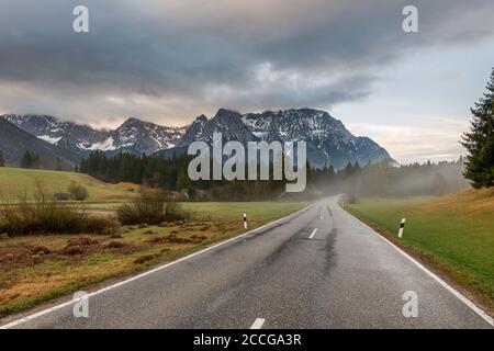 Early morning on the country road at Schmalensee near Mittenwald, the road is wet with rain, light fog rises and the Karwendel towers in the backgroun Stock Photo