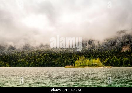 A small green island with a log cabin in the spring-like Eibsee below the cloud-covered Zugspitze. Fresh snow lies on the forest in the background dur Stock Photo