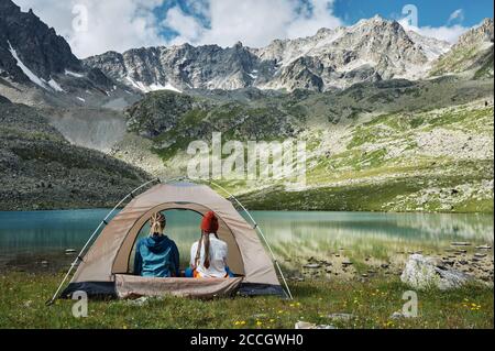Two young women in the tourist tent on the green valley against the turquoise lake and mountain snowy range. Extreme hikers in camp. Trekking lifestyl Stock Photo
