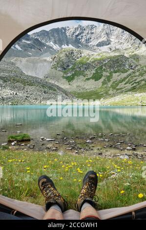 POV selfie shot of mans feet in trekking boots against majestic landscape of mountains and lake. Extreme hiker in tourist tent. Trekking lifestyle. Do Stock Photo