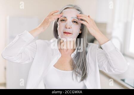 Pretty elderly gray haired woman puts cloth nourishing mask on her face skin for rejuvenating and eliminating defects, while standing on the Stock Photo