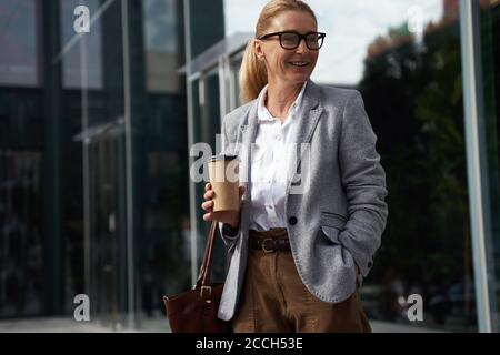 Coffee break. Stylish happy business woman wearing classic eyeglasses holding cup of coffee and smiling while standing in front of glass office Stock Photo