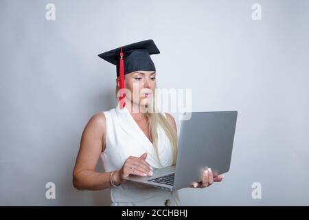 Beautiful blonde graduation cap holds a laptop in her hand, studio light background. Concept, education, learning remotely, online. High quality photo Stock Photo