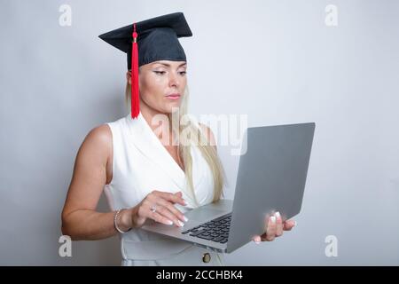 Beautiful blonde graduation cap holds a laptop in her hand, studio light background. Concept, education, learning remotely, online. High quality photo Stock Photo