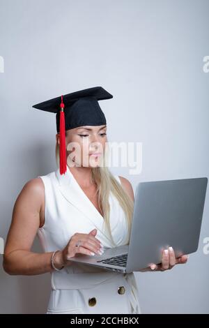 Beautiful blonde graduation cap holds a laptop in her hand, studio light background. Concept, education, learning remotely, online. High quality photo Stock Photo