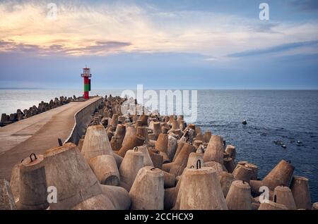 Pier with lighthouse protected by concrete breakwater tetrapods at sunset. Stock Photo