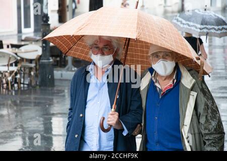 Belgrade, Serbia - August 5, 2020: Two generation elderly men wearing protective face mask walking under umbrella on a rainy  day in the city street Stock Photo