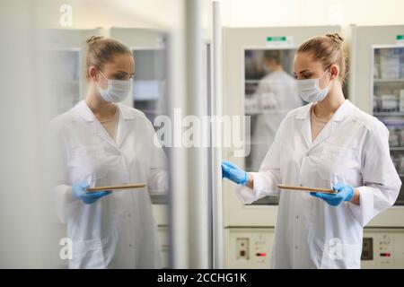 Female doctor in mask and in white coat using digital tablet while working at the laboratory Stock Photo