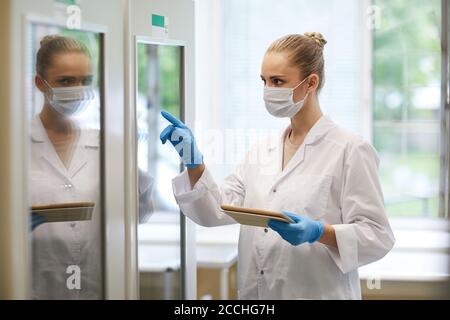 Young female doctor in mask and in white coat using digital tablet at work she pointing at fridge while working at the lab Stock Photo