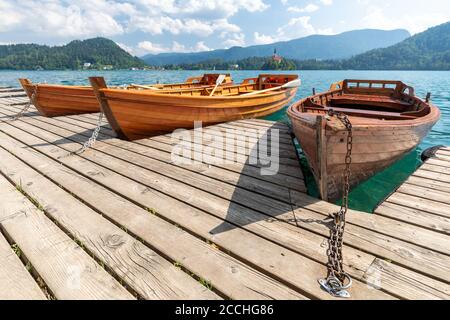 Wide angle view of the Slovenian lake Bled, with a wooden pier and three row boats moored, under a blue summer sky with puffy clouds Stock Photo