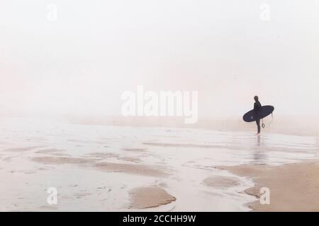 A surfer alone on the beach in sea fog in contemplative mood. Stock Photo