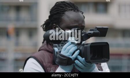 Close up, african black videographer with medikal mask looking through the viewfinder on video camera . High quality photo Stock Photo