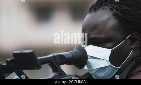 Close up, african black videographer with medikal mask looking through the viewfinder on video camera . High quality photo Stock Photo