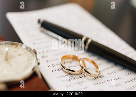 Gold wedding rings on a sheet covered with writing with a black handle and a wrist watch. Stock Photo