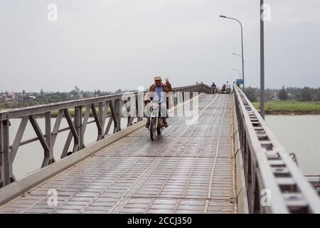 Hoi An / Vietnam - January 18, 2020: Vietnamese man climbs on motorcycle and making victory sign while crossing long bridge Stock Photo