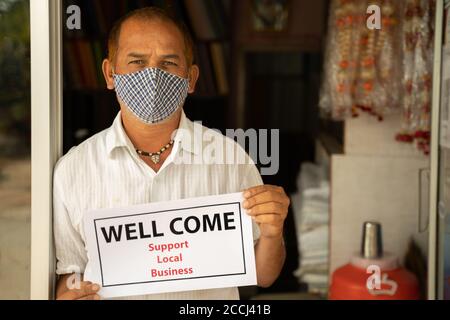 Small business owner in medical mask holding Wellcome notice infront of door after store reopening during coronavirus or covid-19 - concept of support Stock Photo